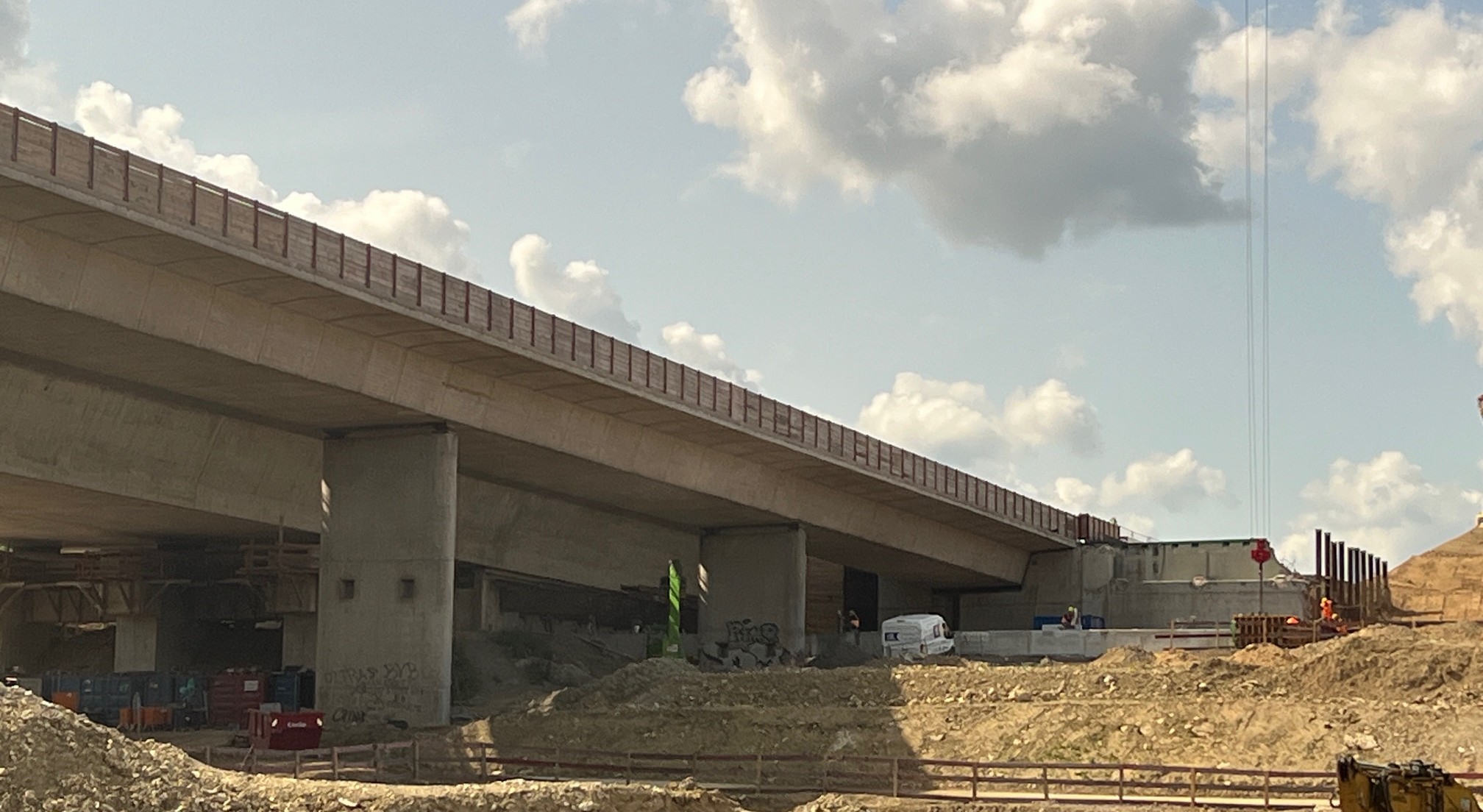 Liedbachtalbruecke - Das Bild zeigt eine Baustelle mit Brückenbauwerk, Betonpfeilern, Baumaschinen, Arbeitern, Geröllhaufen und einem bewölkten Himmel.                                    